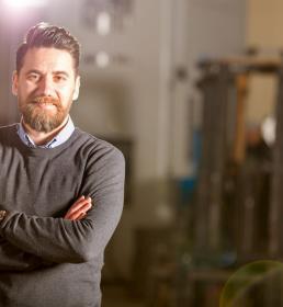 A man in a factory wearing a shirt and jumper smiles at the camera with his arms crossed