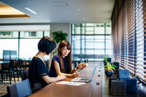 Two women sit by a window talking about a project on the laptop on a table in front of them